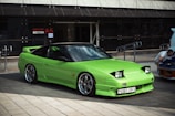 A bright green sports car with a sleek, aerodynamic design is parked outdoors near a modern building with a glass facade. The car features prominent pop-up headlights and shiny alloy wheels. A man is seen working on a blue vehicle in the background.