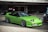 A bright green sports car with a sleek, aerodynamic design is parked outdoors near a modern building with a glass facade. The car features prominent pop-up headlights and shiny alloy wheels. A man is seen working on a blue vehicle in the background.