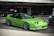 A bright green sports car with a sleek, aerodynamic design is parked outdoors near a modern building with a glass facade. The car features prominent pop-up headlights and shiny alloy wheels. A man is seen working on a blue vehicle in the background.