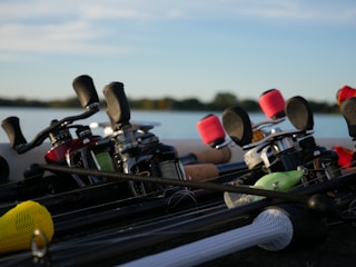 A collection of fishing rods and reels with various colored line spools laying on a surface near a body of water. The background shows part of a lake with a clear sky, and the foreground is dominated by the fishing equipment, including distinctively colored items like a yellow grip and a green lure.