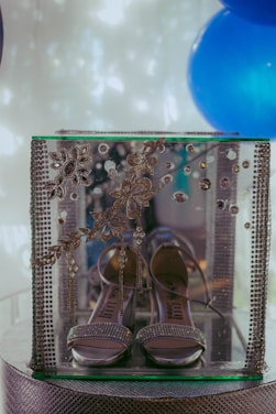 A pair of elegant, high-heeled sandals displayed inside a transparent glass case adorned with intricate, floral and gemstone designs. The case is surrounded by blue decorations and positioned on a textured surface.