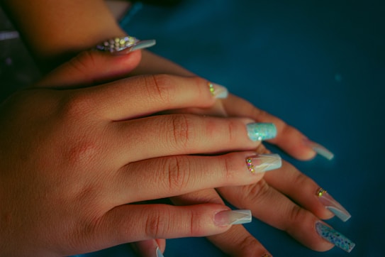 Hands with long, manicured nails displaying various nail art designs including rhinestones and glitter. The nails have a glossy finish, with some featuring sparkly turquoise and others clear with embellishments. The skin tone is warm and contrasts with the blue background.