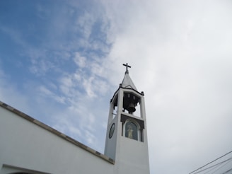 A white church tower with a cross on top and a bell inside, set against a partly cloudy sky. There is a statue visible in a window at the base of the tower.