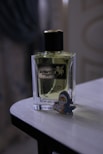 A bottle of perfume labeled 'Aventos' is positioned on a wooden surface. Next to it is a cute cartoon figure shaped like a baby wearing a shark costume.