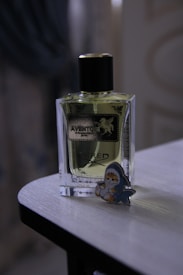 A bottle of perfume labeled 'Aventos' is positioned on a wooden surface. Next to it is a cute cartoon figure shaped like a baby wearing a shark costume.