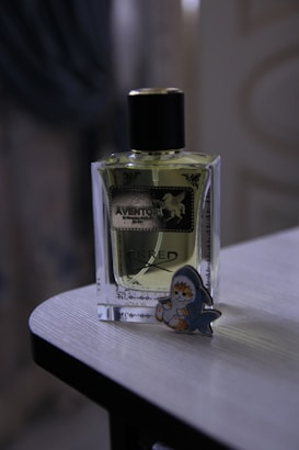 A bottle of perfume labeled 'Aventos' is positioned on a wooden surface. Next to it is a cute cartoon figure shaped like a baby wearing a shark costume.