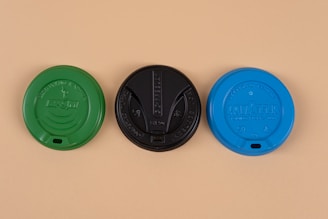 Three coffee cup lids are arranged side by side, each featuring different colors and brand insignia. The left lid is green, the center lid is black, and the right lid is blue, all set against a beige background.