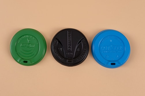 Three coffee cup lids are arranged side by side, each featuring different colors and brand insignia. The left lid is green, the center lid is black, and the right lid is blue, all set against a beige background.