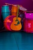 A collection of musical instruments including a teal conga, a vibrant orange djembe, a wooden acoustic guitar, and a large pink drum, all set against a deep purple background. The instruments are arranged closely together, showcasing a variety of textures and materials.