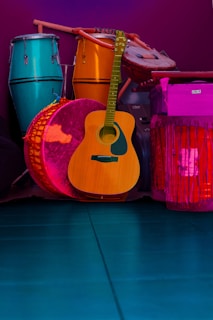 A collection of musical instruments including a teal conga, a vibrant orange djembe, a wooden acoustic guitar, and a large pink drum, all set against a deep purple background. The instruments are arranged closely together, showcasing a variety of textures and materials.