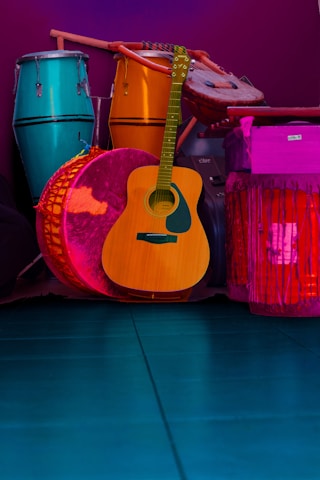 A collection of musical instruments including a teal conga, a vibrant orange djembe, a wooden acoustic guitar, and a large pink drum, all set against a deep purple background. The instruments are arranged closely together, showcasing a variety of textures and materials.