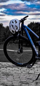A bicycle with a white helmet resting on the handlebars is positioned on a cobblestone path. The bicycle is predominantly blue with a dark frame and has a rugged, sporty design. In the background, trees are visible under a sky painted with clouds in hues of blue, purple, and white, suggesting a serene outdoor setting.