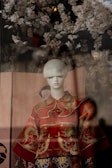 A mannequin dressed in an ornate red garment featuring intricate gold patterns and dragon motifs. The background is adorned with delicate cherry blossom branches, creating a soft and cultural ambiance.