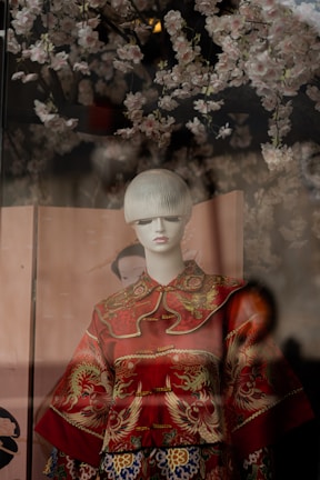 A mannequin dressed in an ornate red garment featuring intricate gold patterns and dragon motifs. The background is adorned with delicate cherry blossom branches, creating a soft and cultural ambiance.