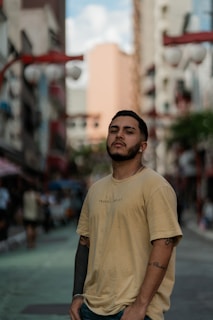 A young man is standing confidently in an urban street setting. The background features blurred city buildings and traditional lanterns, suggesting a vibrant urban neighborhood.
