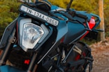 A close-up view of a motorcycle with a visible front headlight and number plate. The bike has a sleek design with black and red accents, sturdy handlebars, and a rugged build. The surroundings are out of focus, showing a hint of greenery in the background.