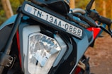 Close-up view of a motorcycle's front headlight and license plate. The license plate displays 'TN 13AA 0868' and is mounted above the headlight, which features a clear, angular design. The handlebars and body of the motorcycle are visible, with a combination of black and red colors.