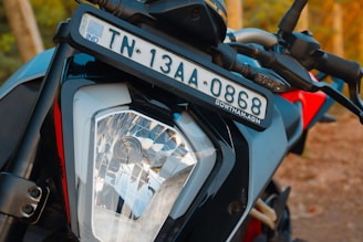 Close-up view of a motorcycle's front headlight and license plate. The license plate displays 'TN 13AA 0868' and is mounted above the headlight, which features a clear, angular design. The handlebars and body of the motorcycle are visible, with a combination of black and red colors.