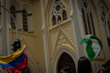 A large beige cathedral with intricate gothic architecture, featuring tall arched windows and decorative carvings. In front of the building, people are holding two large flags, one with yellow, blue, and red stripes and another with green and white colors. The scene is lively and dynamic, suggesting a gathering or event.