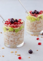 Two glass cups filled with a mixture of granola and milk, topped with fresh fruits, which include pomegranate seeds, blueberries, and slices of kiwi and banana. Stainless steel spoons are placed in each cup, and a few pieces of granola and fruits are scattered around on the light-colored surface.
