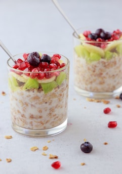 Two glass cups filled with a mixture of granola and milk, topped with fresh fruits, which include pomegranate seeds, blueberries, and slices of kiwi and banana. Stainless steel spoons are placed in each cup, and a few pieces of granola and fruits are scattered around on the light-colored surface.