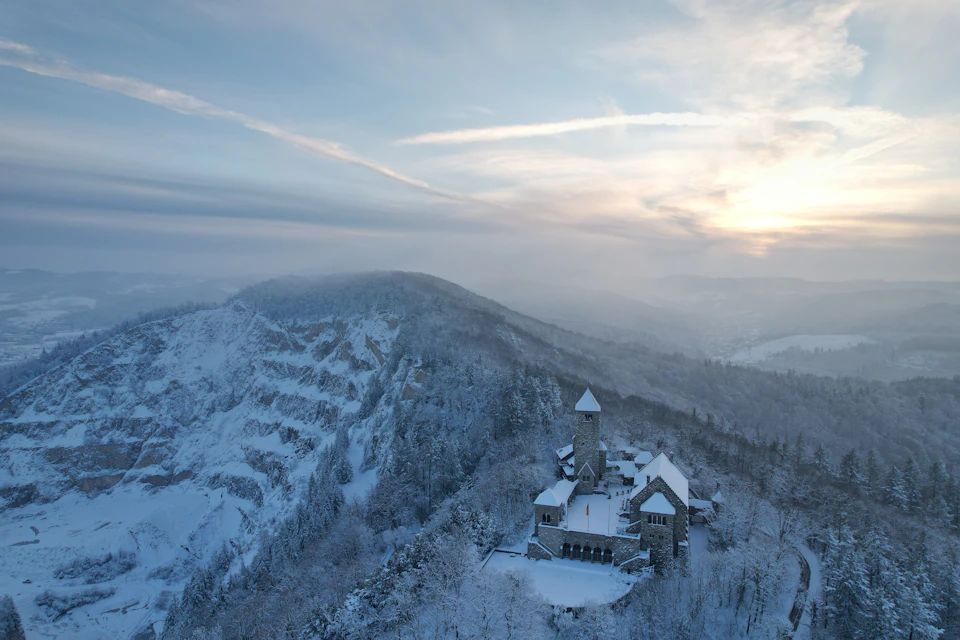 The image presents an aerial view of a serene winter landscape, dominated by a large mountain blanketed in snow with a small church perched on its peak. The church features a tall tower and a steeple, adding to its majestic presence against the backdrop of the snowy mountain range. The sky above is a clear blue, dotted with wispy clouds that cast soft shadows over the scene below.