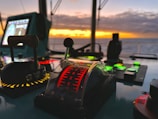 A control panel on a ship's bridge with various buttons and levers, illuminated against a backdrop of a striking sunset over the ocean. The panel includes a throttle lever with increments marked in red and surrounding lights in different colors.