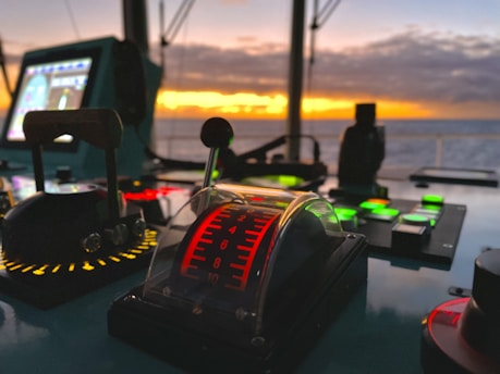 A control panel on a ship's bridge with various buttons and levers, illuminated against a backdrop of a striking sunset over the ocean. The panel includes a throttle lever with increments marked in red and surrounding lights in different colors.
