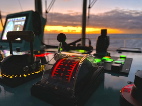 A control panel on a ship's bridge with various buttons and levers, illuminated against a backdrop of a striking sunset over the ocean. The panel includes a throttle lever with increments marked in red and surrounding lights in different colors.