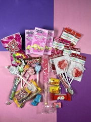 A colorful assortment of candies including various lollipops, individually wrapped candies, chocolate bars, conversation hearts, and gummy candies, all arranged on a split purple and pink background.