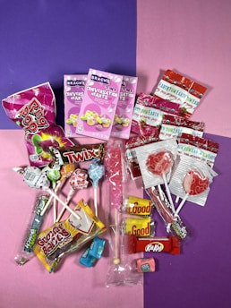 A colorful assortment of candies including various lollipops, individually wrapped candies, chocolate bars, conversation hearts, and gummy candies, all arranged on a split purple and pink background.