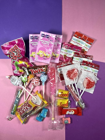 A colorful assortment of candies including various lollipops, individually wrapped candies, chocolate bars, conversation hearts, and gummy candies, all arranged on a split purple and pink background.