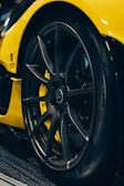 Close-up view of a sleek, black car tire with intricate alloy wheel design, set against a vibrant yellow car body. The surface of the tire appears glossy and well-maintained, with lighting highlighting the modern wheel structure.