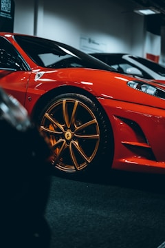 A close-up view of a luxurious red sports car with gold rims, parked indoors. The sleek body design and glossy finish emphasize its high-end nature.