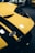 A close-up of the front section of a yellow sports car featuring black stripes and a badge with the Ford logo. The car is likely positioned in a showroom or garage with other cars visible in the blurred background.