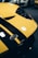 A close-up of the front section of a yellow sports car featuring black stripes and a badge with the Ford logo. The car is likely positioned in a showroom or garage with other cars visible in the blurred background.