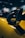 A close-up shot of a car's side mirror with a glossy, carbon fiber design and the letters 'P1' in yellow. The background features soft-focus shapes, enhancing the emphasis on the sleek and stylish component.