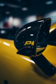 A close-up shot of a car's side mirror with a glossy, carbon fiber design and the letters 'P1' in yellow. The background features soft-focus shapes, enhancing the emphasis on the sleek and stylish component.