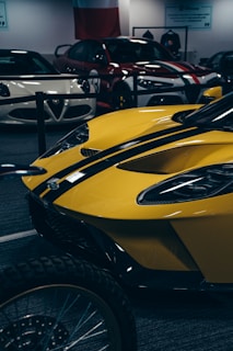 A collection of sports cars is on display in an indoor showroom. The foreground features a striking yellow sports car with black racing stripes. Behind it, other sleek vehicles, including white and red models, are positioned. The environment is sophisticated, emphasizing vehicle design and allure.