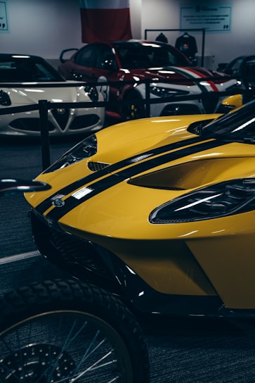 A collection of sports cars is on display in an indoor showroom. The foreground features a striking yellow sports car with black racing stripes. Behind it, other sleek vehicles, including white and red models, are positioned. The environment is sophisticated, emphasizing vehicle design and allure.