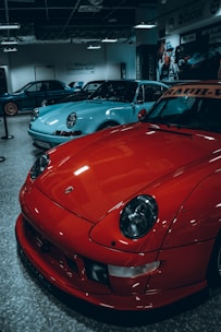 A collection of classic sports cars displayed indoors, with a prominent red car in the foreground and two additional cars, one blue and one black, visible in the background. The setting is likely a showroom or museum with dim lighting and various automotive posters and signage on the walls.