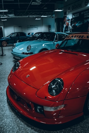 A collection of classic sports cars displayed indoors, with a prominent red car in the foreground and two additional cars, one blue and one black, visible in the background. The setting is likely a showroom or museum with dim lighting and various automotive posters and signage on the walls.