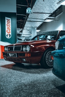 A red vintage car is parked indoors under a metal ceiling with exposed pipes and ductwork. The foreground reveals the front half of a polished teal car. A Castrol sign is mounted on the wall behind the red car. The area has a commercial or industrial appearance, highlighted by the speckled flooring and emergency exit sign.