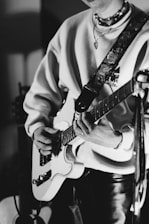A person playing an electric guitar, wearing a cozy sweater and layered necklaces. The focus is on the hands and guitar, with a floral-patterned guitar strap. The monochrome image adds a classic and artistic feel.