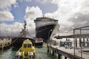 A large cruise ship is docked at a port with several smaller tugboats surrounding it. The scene features a bustling pier, and the sky is partly cloudy, creating an active and busy maritime setting.