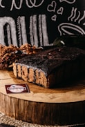 A rich chocolate cake sits on a wooden slab, surrounded by rustic decorations and chalkboard-style illustrations of food and hearts in the background. A label reading 'Delicias da Nila' is displayed in the foreground.