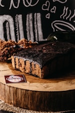 A rich chocolate cake sits on a wooden slab, surrounded by rustic decorations and chalkboard-style illustrations of food and hearts in the background. A label reading 'Delicias da Nila' is displayed in the foreground.