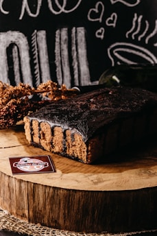 A rich chocolate cake sits on a wooden slab, surrounded by rustic decorations and chalkboard-style illustrations of food and hearts in the background. A label reading 'Delicias da Nila' is displayed in the foreground.