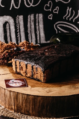 A rich chocolate cake sits on a wooden slab, surrounded by rustic decorations and chalkboard-style illustrations of food and hearts in the background. A label reading 'Delicias da Nila' is displayed in the foreground.