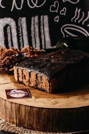 A rich chocolate cake sits on a wooden slab, surrounded by rustic decorations and chalkboard-style illustrations of food and hearts in the background. A label reading 'Delicias da Nila' is displayed in the foreground.